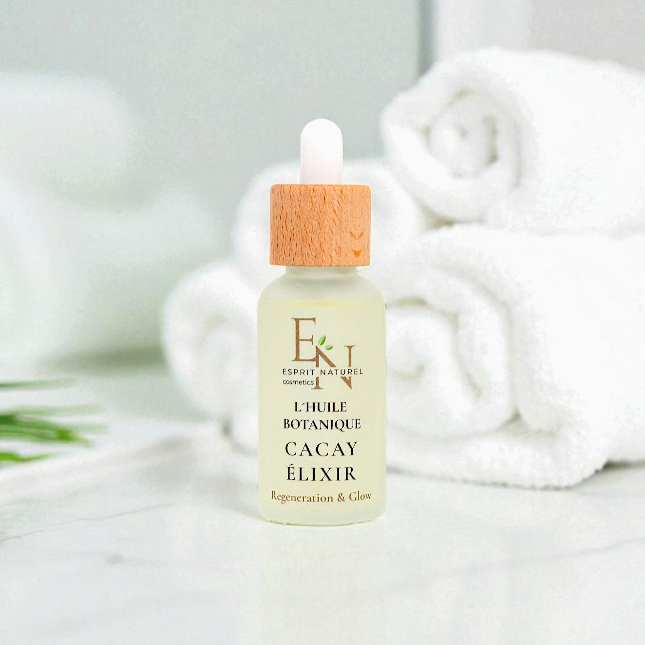 Esprit Naturel l´huile Botanique Bio-Cacayelizir in eleganter Glasflasche – reich an Vitamin A & E, regenerierendes und glättendes Naturöl aus dem Amazonas für Gesicht und Körper. Im Hintergund  Badezimmer, mit weißen Handtüchern