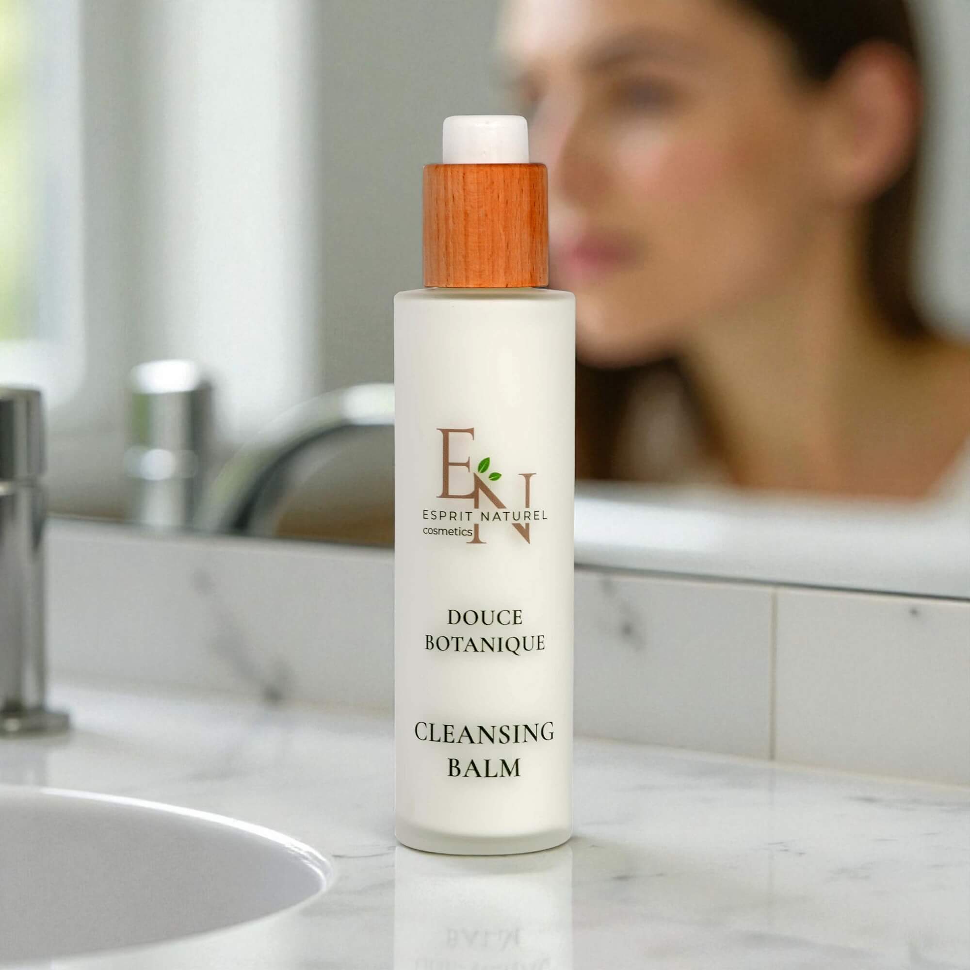 Esprit Naturel Douce Botanique Cleansing Balm auf einem Badezimmerwaschbecken mit verschwommener Frau im Hintergrund – luxuriöse, sanfte Naturkosmetik für die tägliche Gesichtsreinigung.