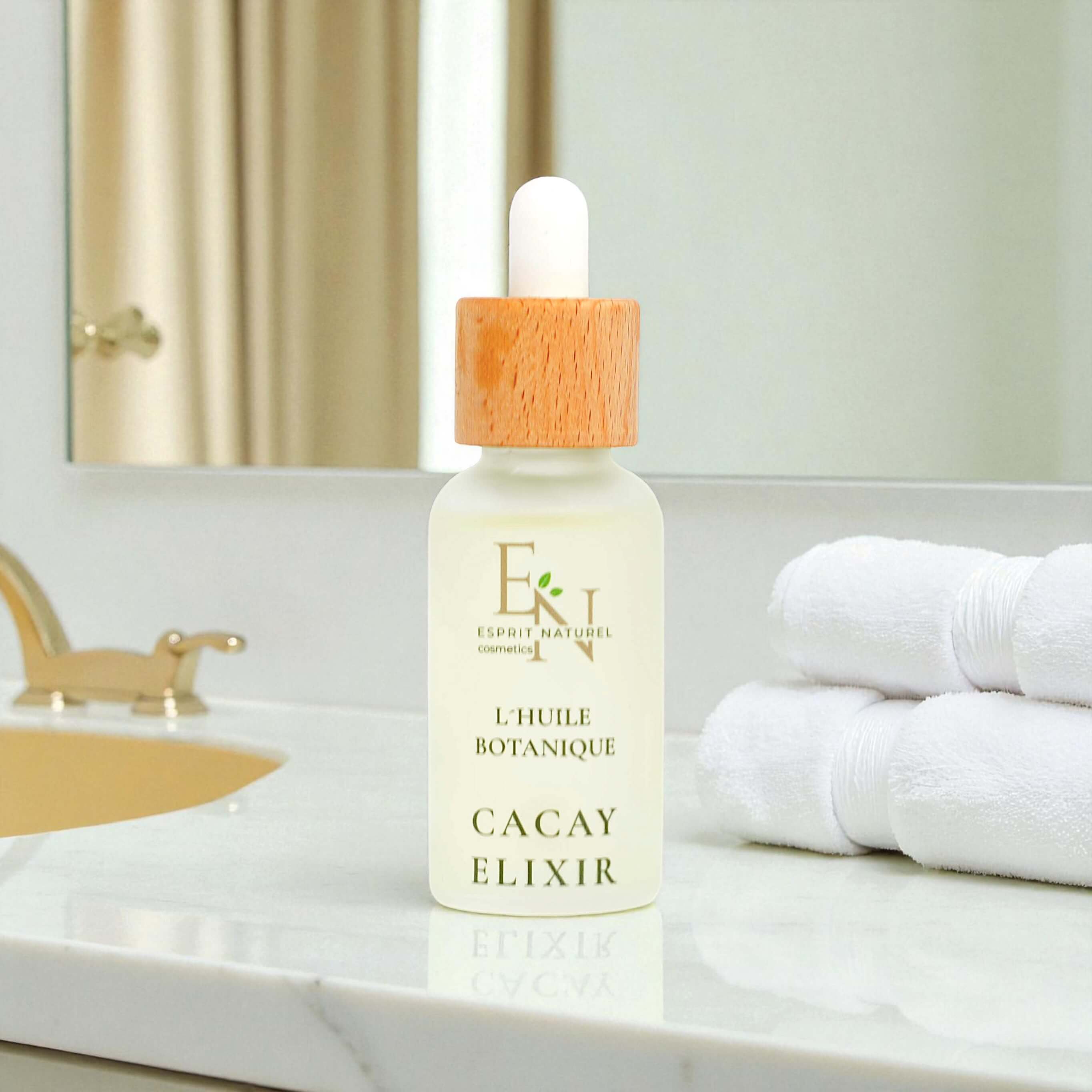 Esprit Naturel l´huile Botanique Bio-Cacayelizir in eleganter Glasflasche – reich an Vitamin A & E, regenerierendes und glättendes Naturöl aus dem Amazonas für Gesicht und Körper. Im Hintergund  Badezimmer, mit weißen Handtüchern